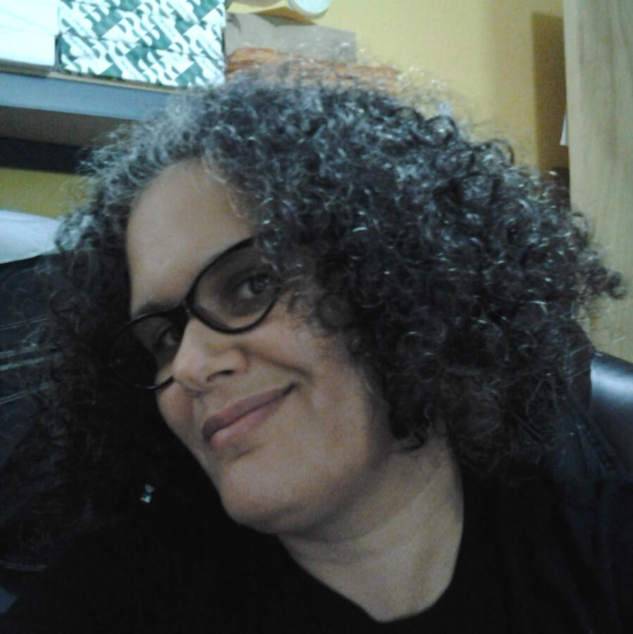 Real Hair Mixed Boricua person with black curly hair and glasses, black shirt and a yellow background.