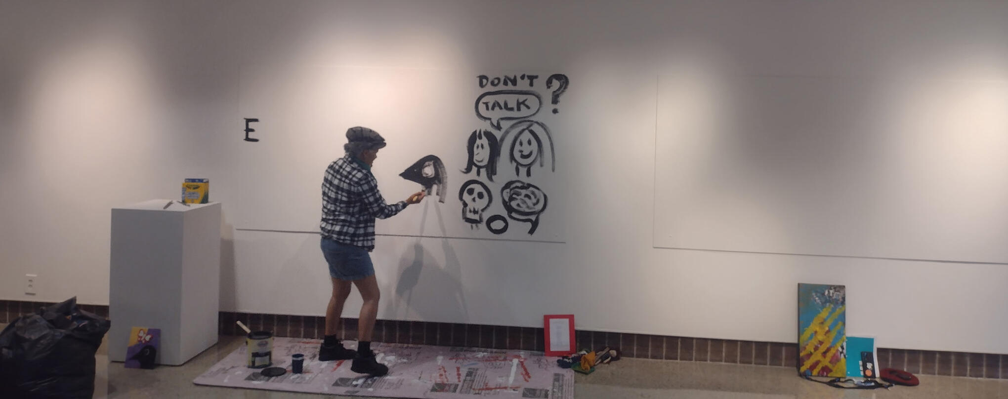 Image by Kelly Travis, courtesy of Lyricality Tanamá dressed in flannel, shorts, a news boy hat, and shoes, painting the Color Me Safe wall at Tanamá Kao's Battle For Safety